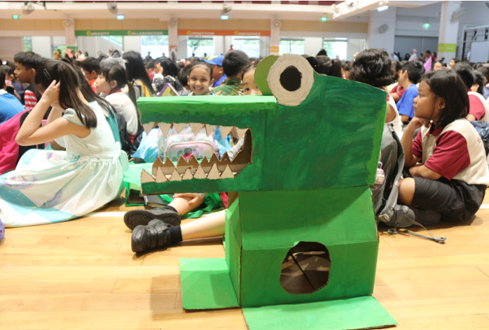 A green cardboard crocodile sculpture sits on a gym floor surrounded by many children seated in a large hall.
