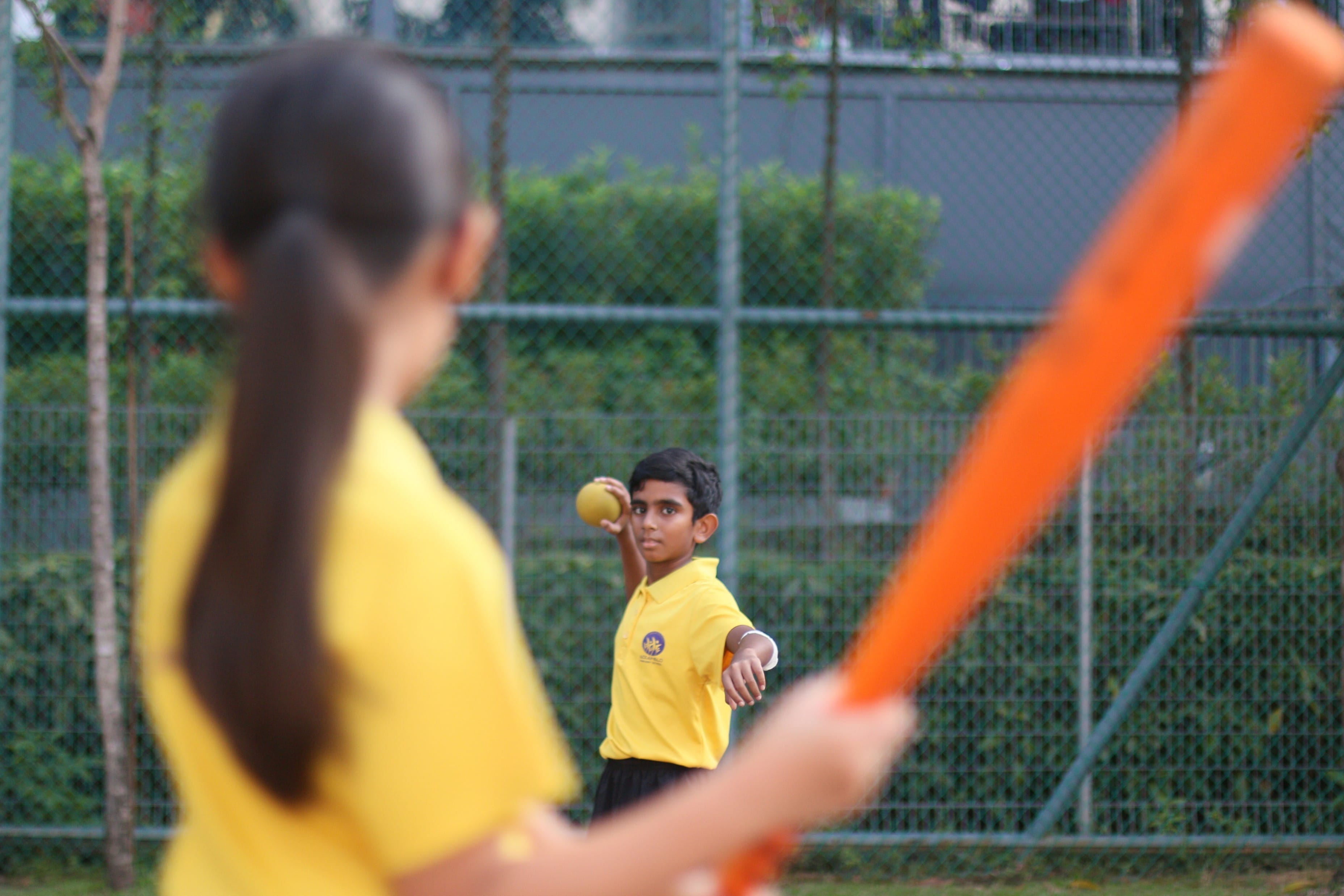 A boy in a yellow shirt prepares to throw a yellow ball toward a girl holding an orange bat outdoors.