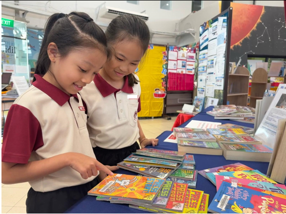 Two young girls in school uniforms smile while browsing colorful books spread on a blue table.