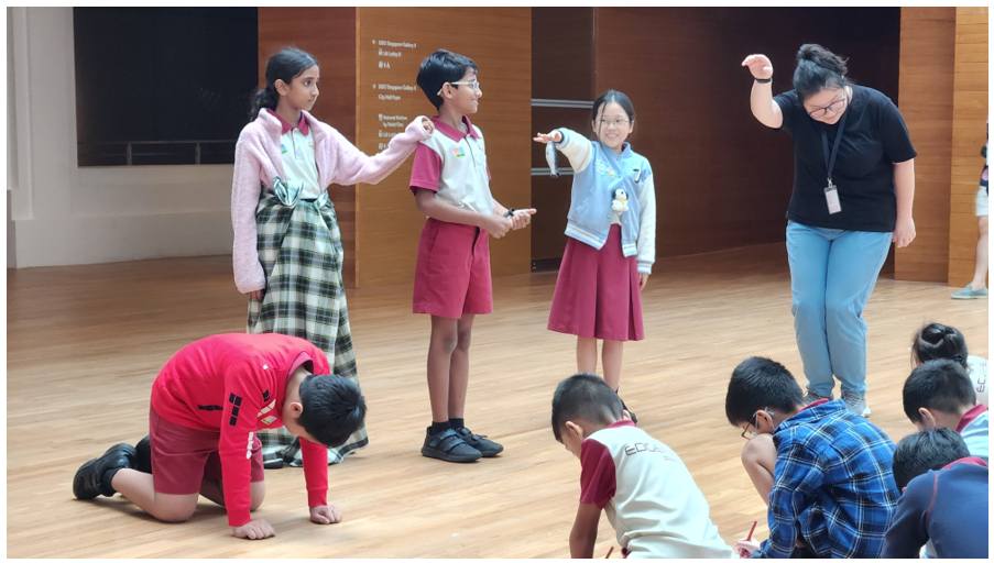 Children in school uniforms participate in a group activity on a wooden floor, guided by an adult with a lanyard.