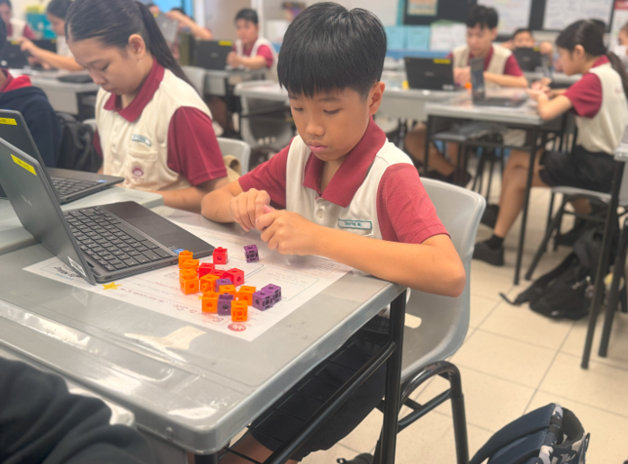 A young student in a school uniform arranges colorful plastic cubes on a desk beside a laptop in a classroom.
