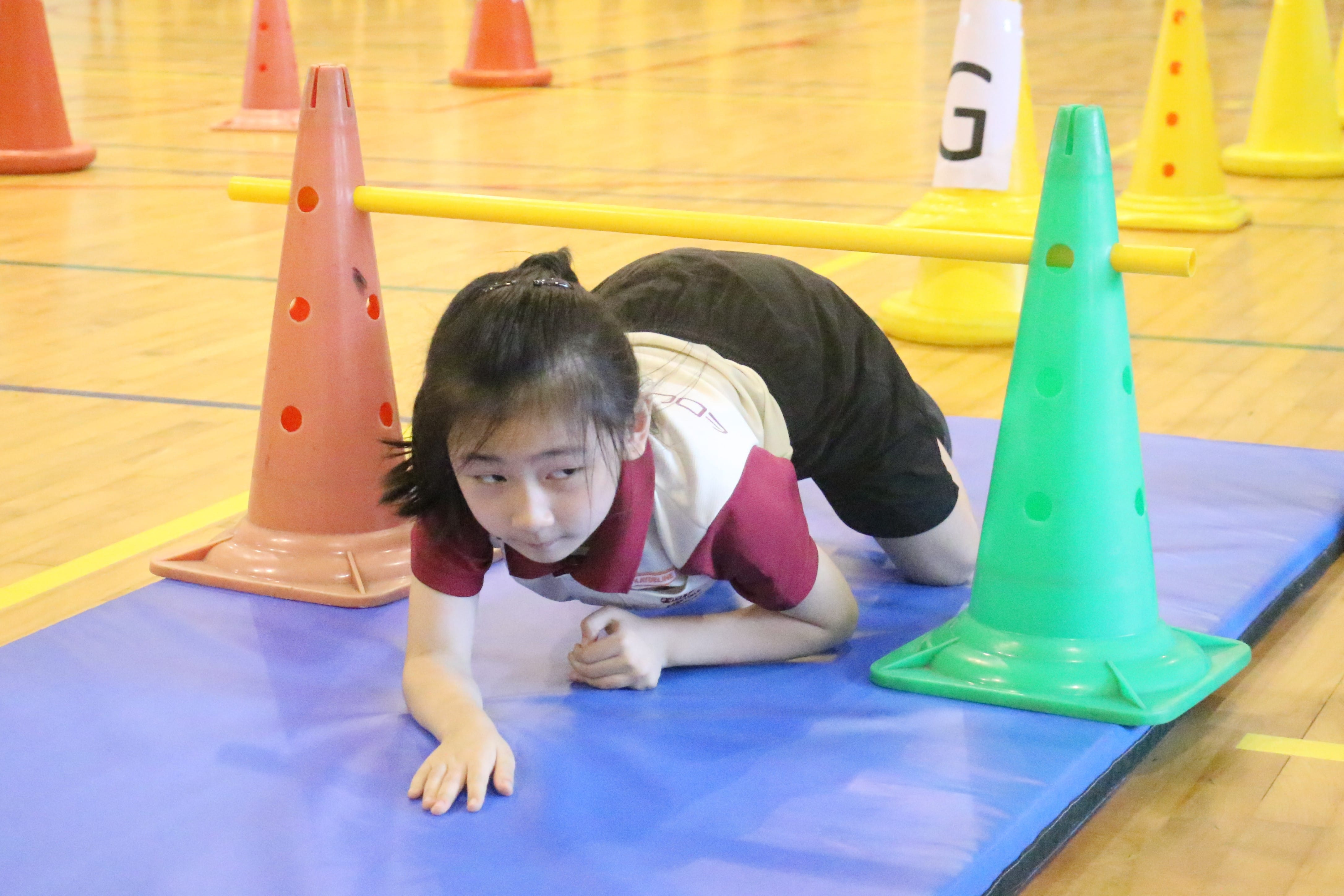 A young girl crawls on a blue mat between colorful cones during an indoor gym obstacle course activity.