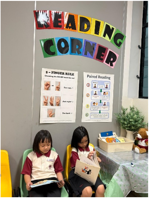Two young students in uniforms sit reading books under a colorful "Reading Corner" sign with posters on the wall.