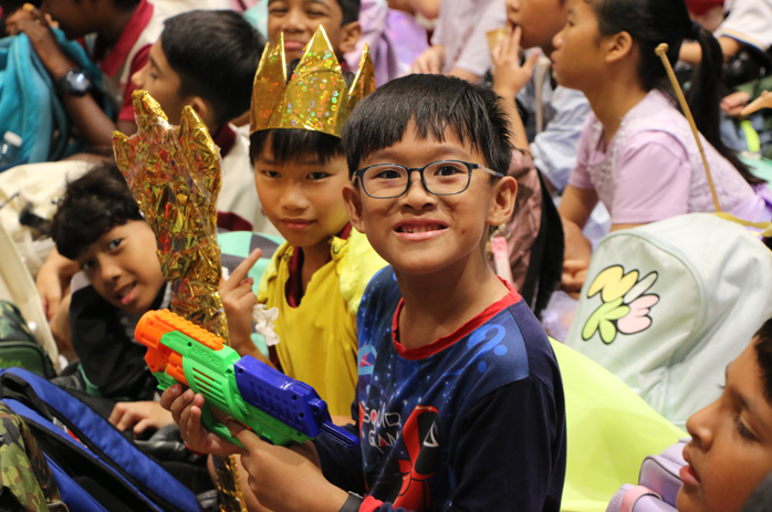 A smiling boy with glasses holds a toy gun and sparkly prop, surrounded by children at a festive gathering.