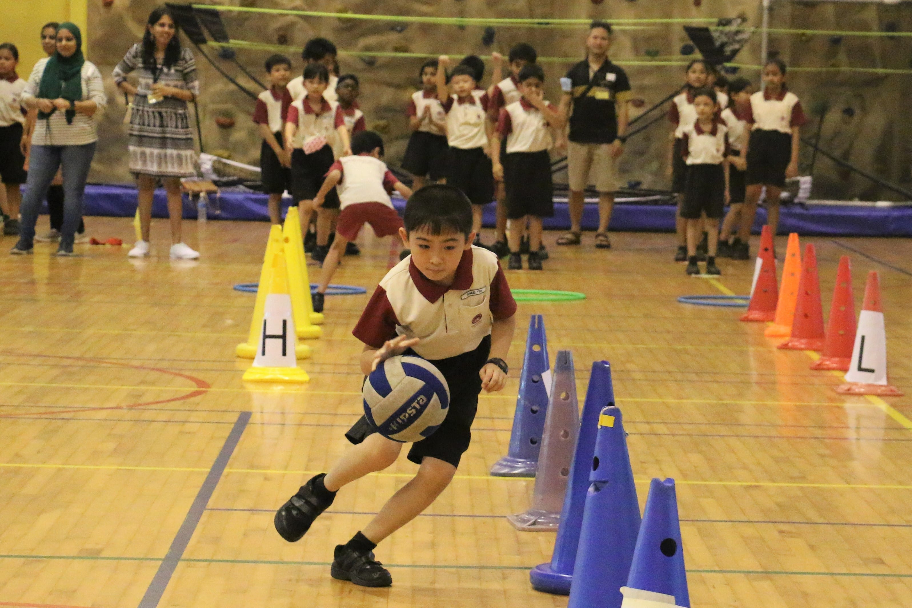 A young boy in school uniform runs with a volleyball through colored cones in a gym while classmates watch.