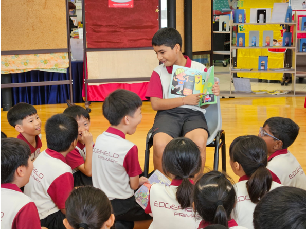A boy sits on a chair reading a colorful picture book aloud to a group of young students seated on the floor.