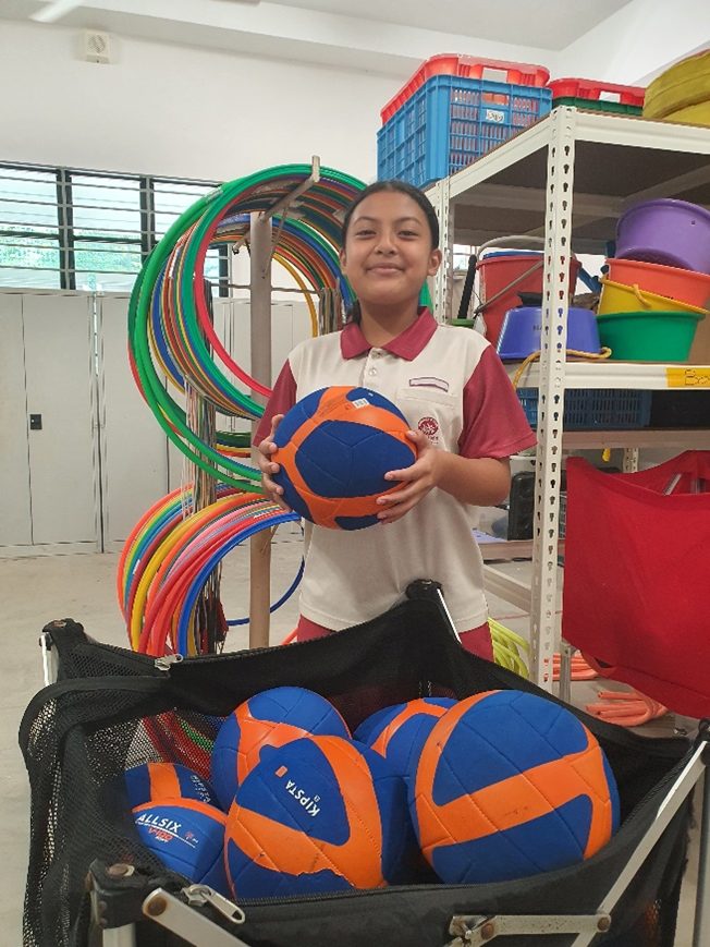 A young girl in a school uniform holds a blue and orange volleyball near a cart of balls and colorful hula hoops.