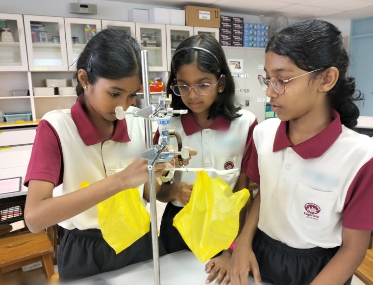 Three school girls in uniforms conduct a science experiment with a stand and yellow plastic bags in a classroom lab.
