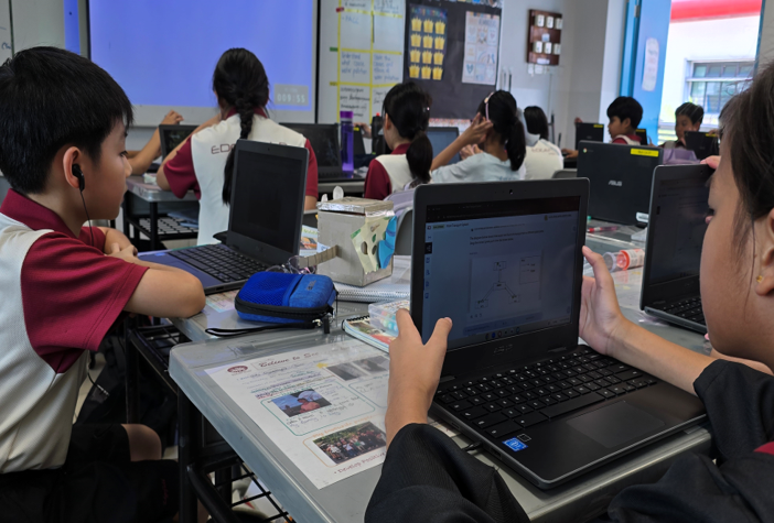 Students in school uniforms use laptops and tablets at desks in a busy classroom.