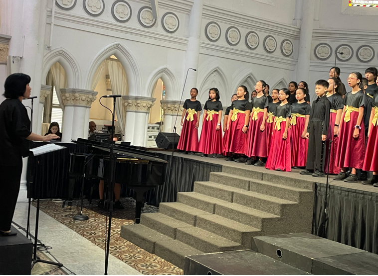 A conductor leads a children's choir in black tops, red skirts, and gold bows on church steps.