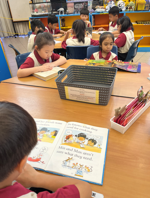 Young children in uniforms read books at tables in a colorful classroom library setting.