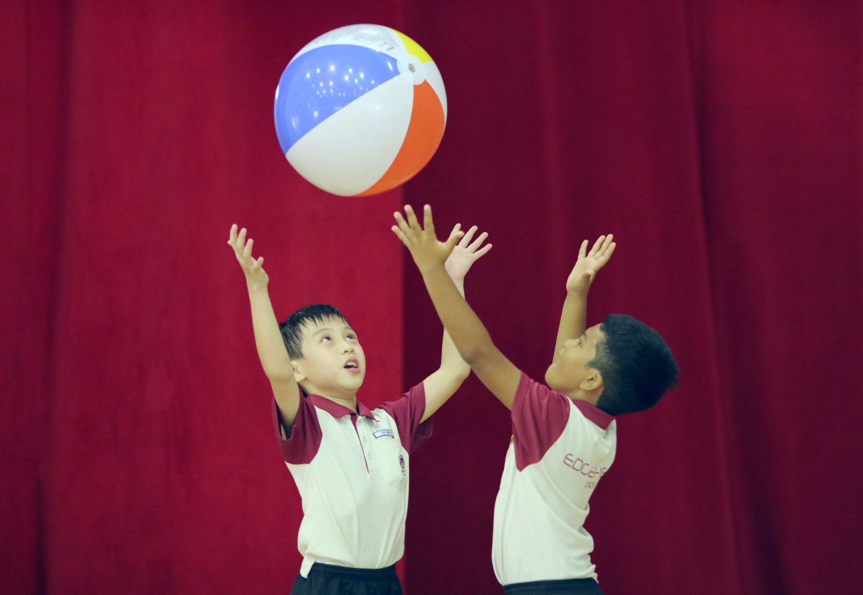 Two boys in school uniforms reach up toward a colorful beach ball against a red curtain backdrop.