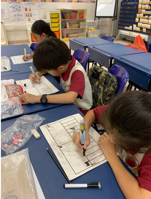 Three young students sit at blue desks in a classroom, writing on paper worksheets with markers.