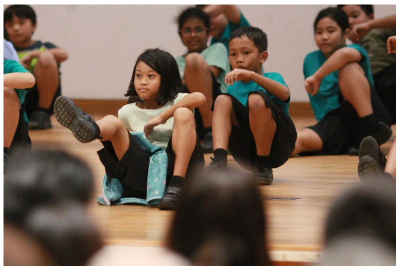Several children sit on a wooden gym floor, most wearing teal shirts, attentively watching something off-camera.