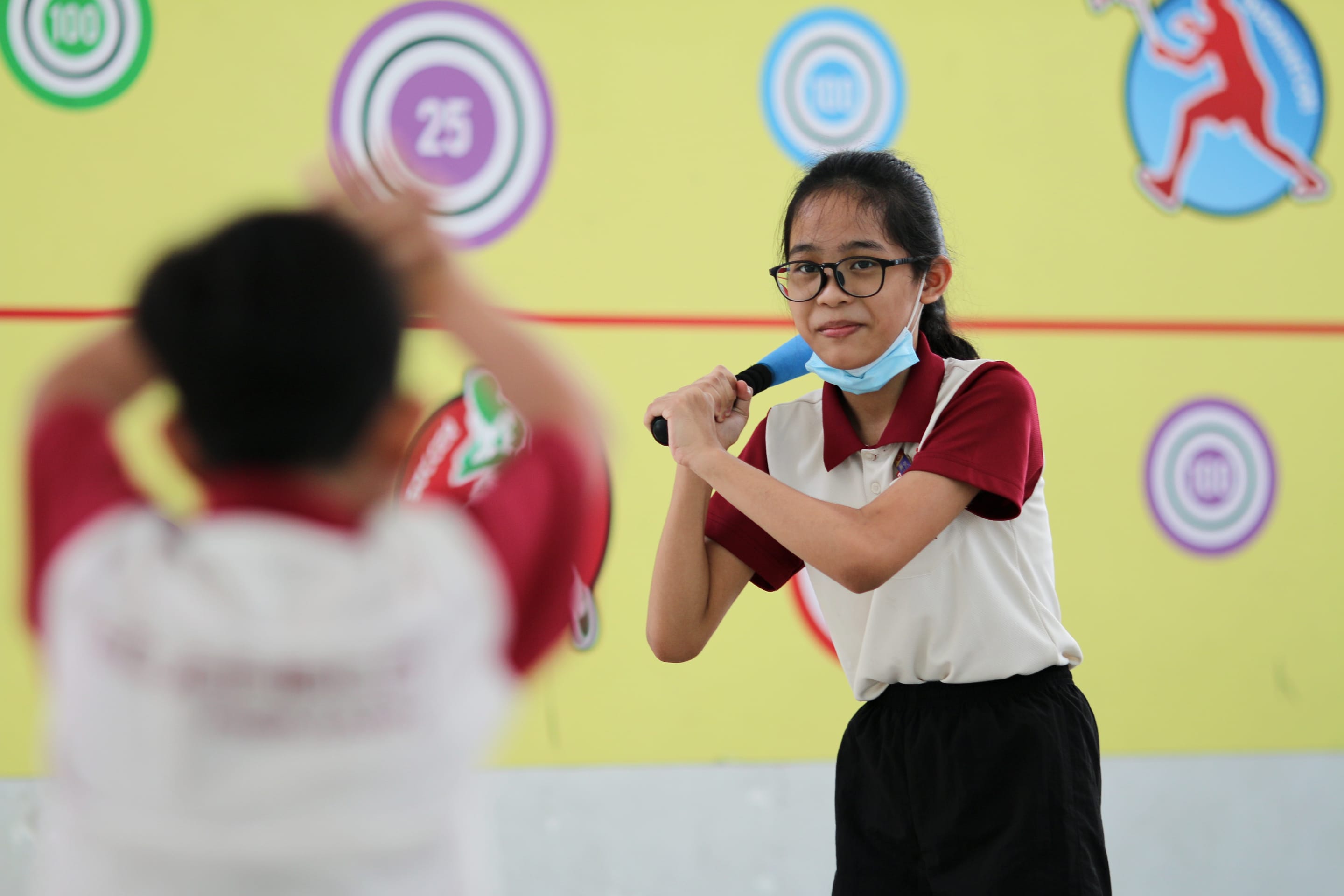 Girl in glasses holds a blue bat ready to swing; another child blurred in foreground; target circles on wall.