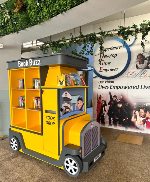 Yellow school bus-shaped bookshelf labeled Book Buzz with a Book Drop slot, filled with children's books indoors.