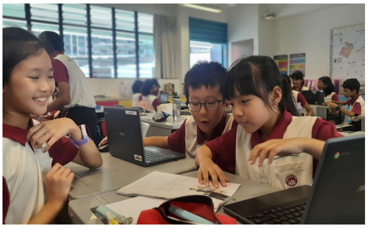 Three young students in school uniforms collaborate around laptops at a classroom desk.