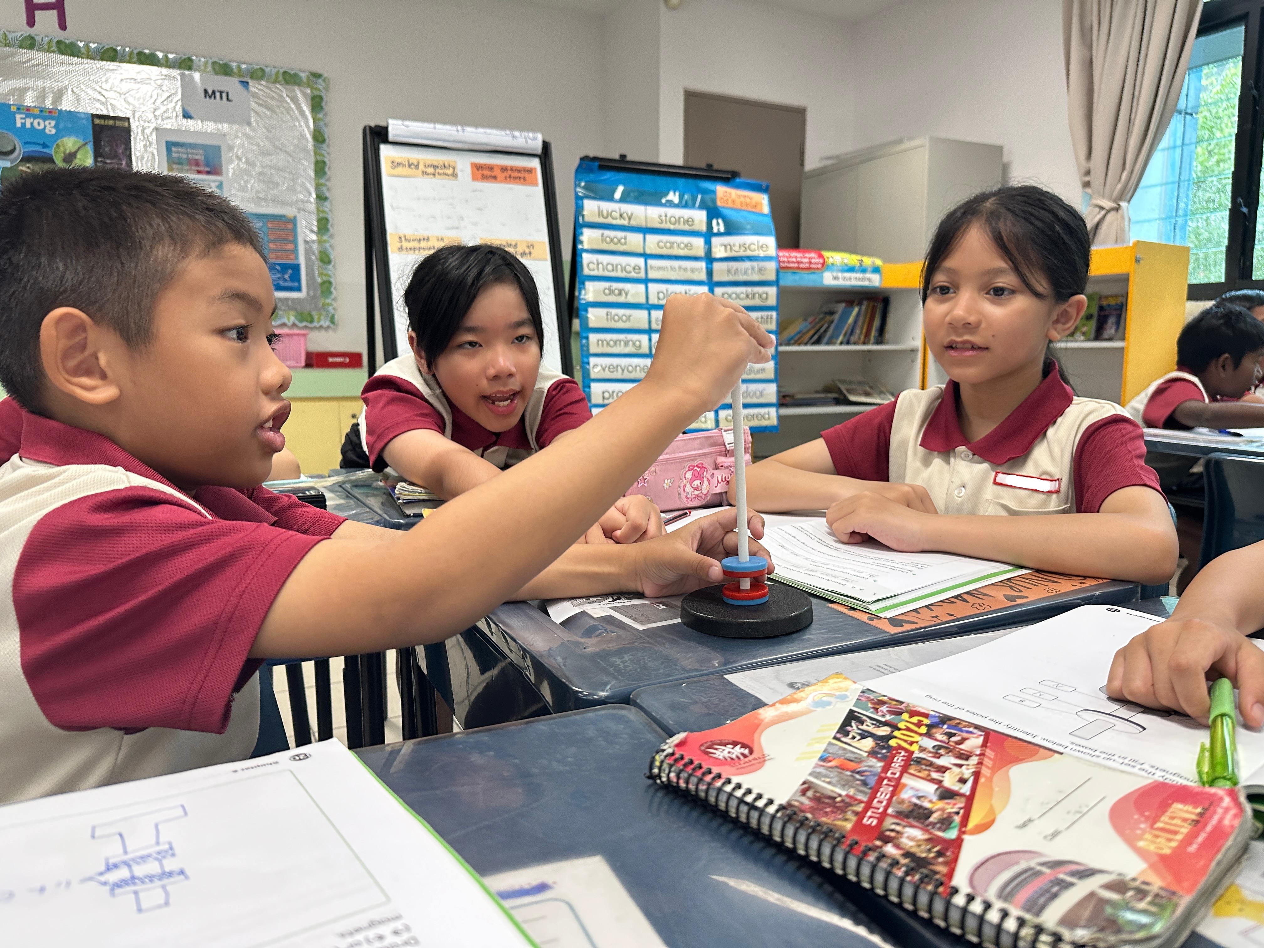 Three young students in uniforms work together at a desk, with a boy spinning a small top during a classroom activity.
