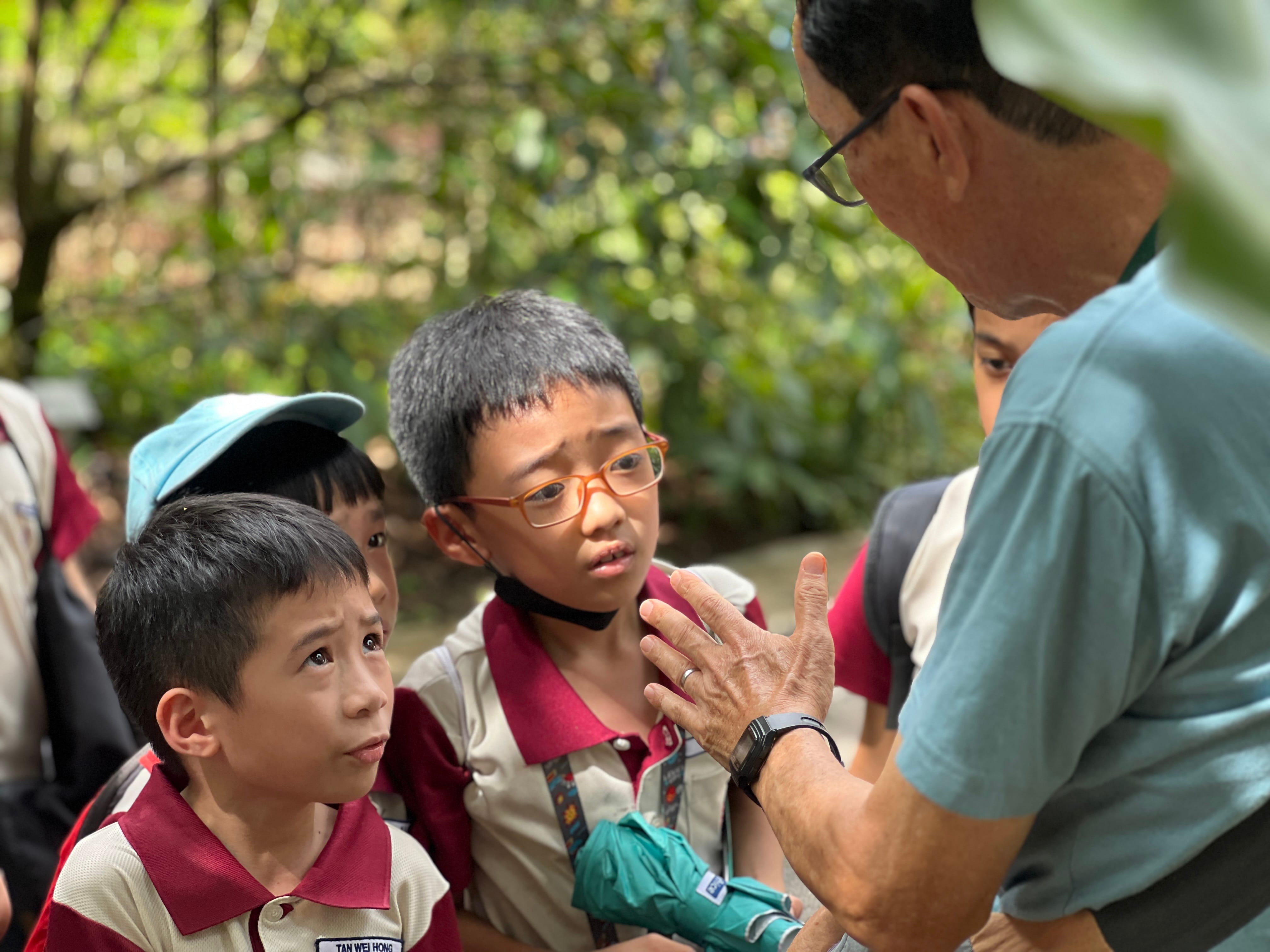 An older man with glasses gestures while speaking to attentive young students in matching school uniforms outdoors.