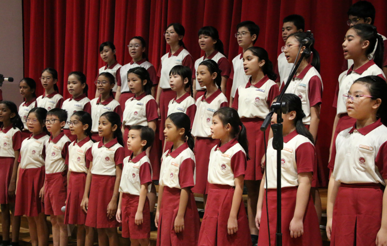 Children in matching red and white uniforms stand in rows on a stage, singing in a choir performance.