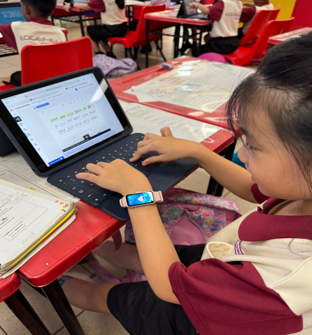 Young girl wearing a smartwatch typing on a tablet with keyboard at a red school desk in a classroom.