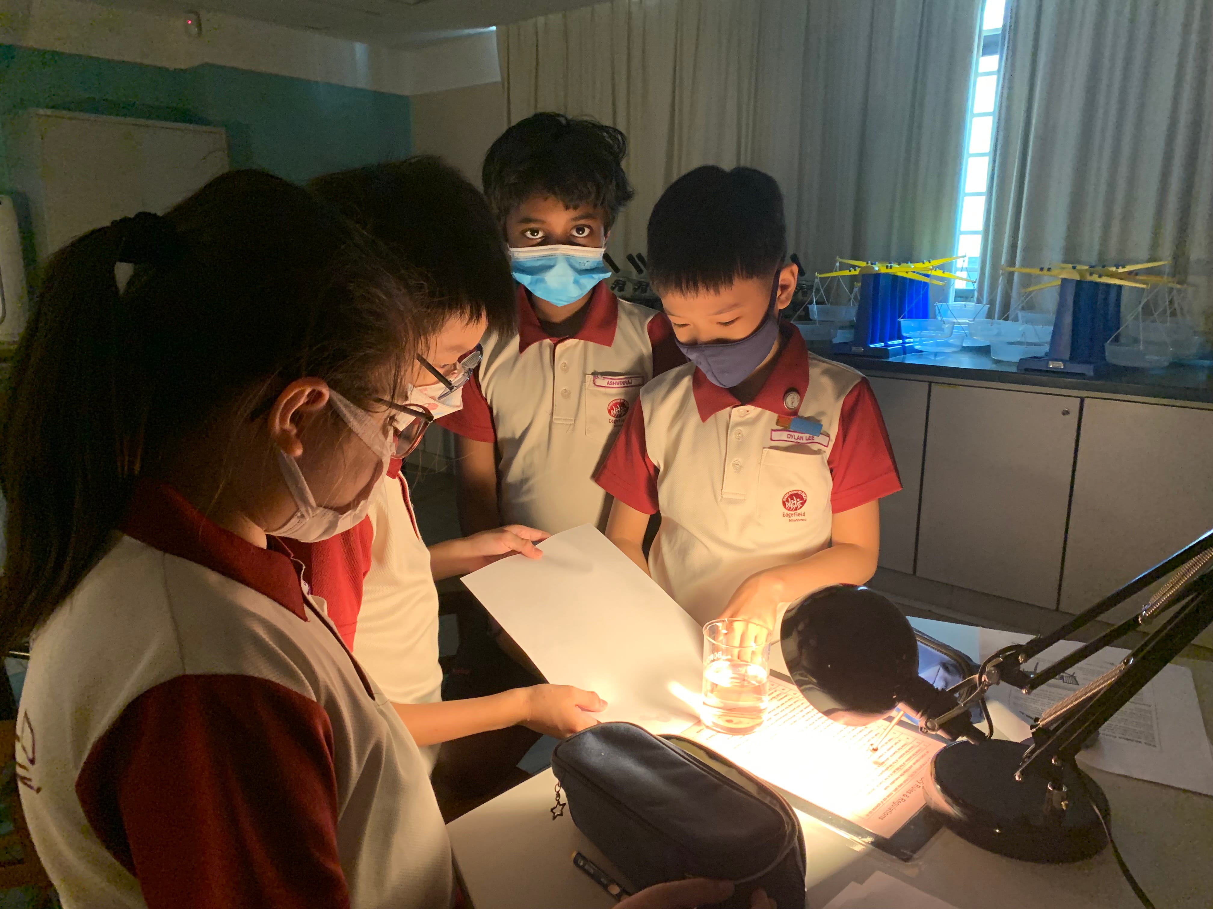 Three masked students in school uniforms gather around a bright desk lamp with a glass of water in a dark classroom.