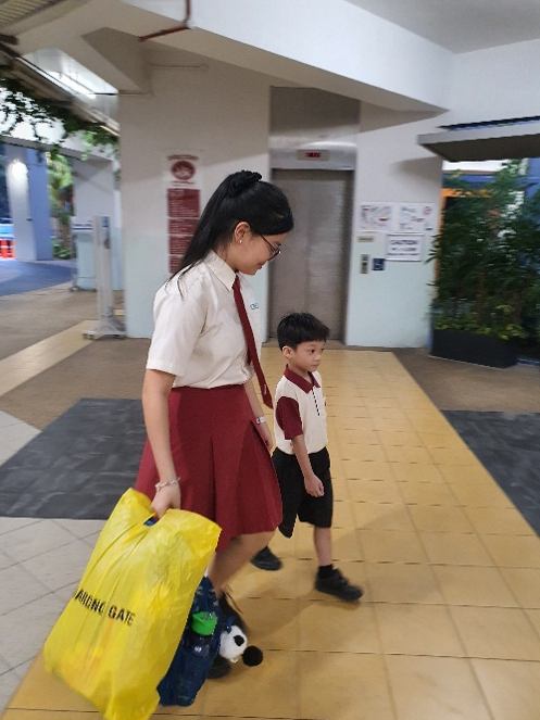 Two children in matching red and white school uniforms walk together, with the older girl carrying a yellow shopping bag.