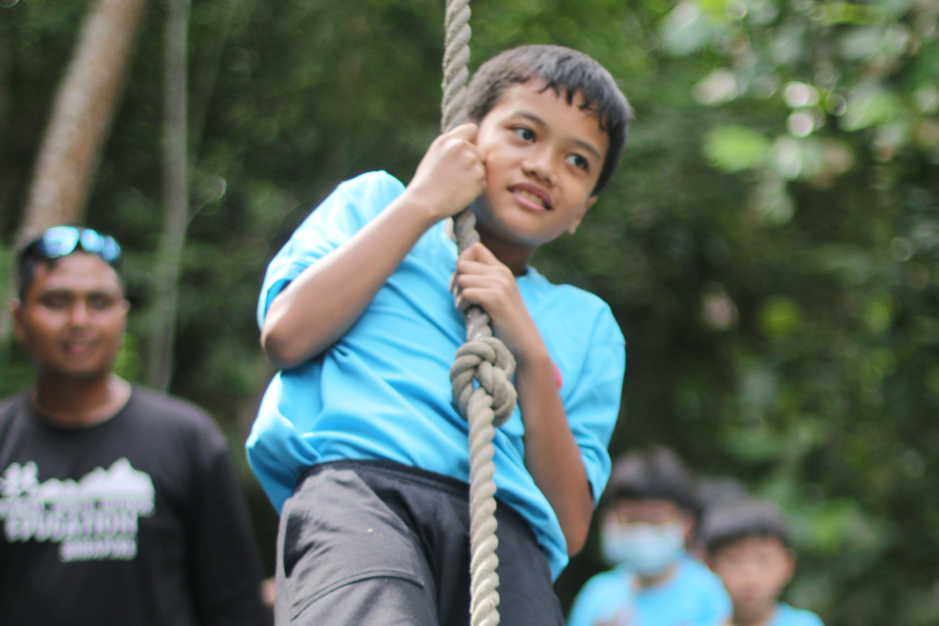 A boy in a blue shirt climbs a knotted rope outdoors, with adults and trees visible in the background.