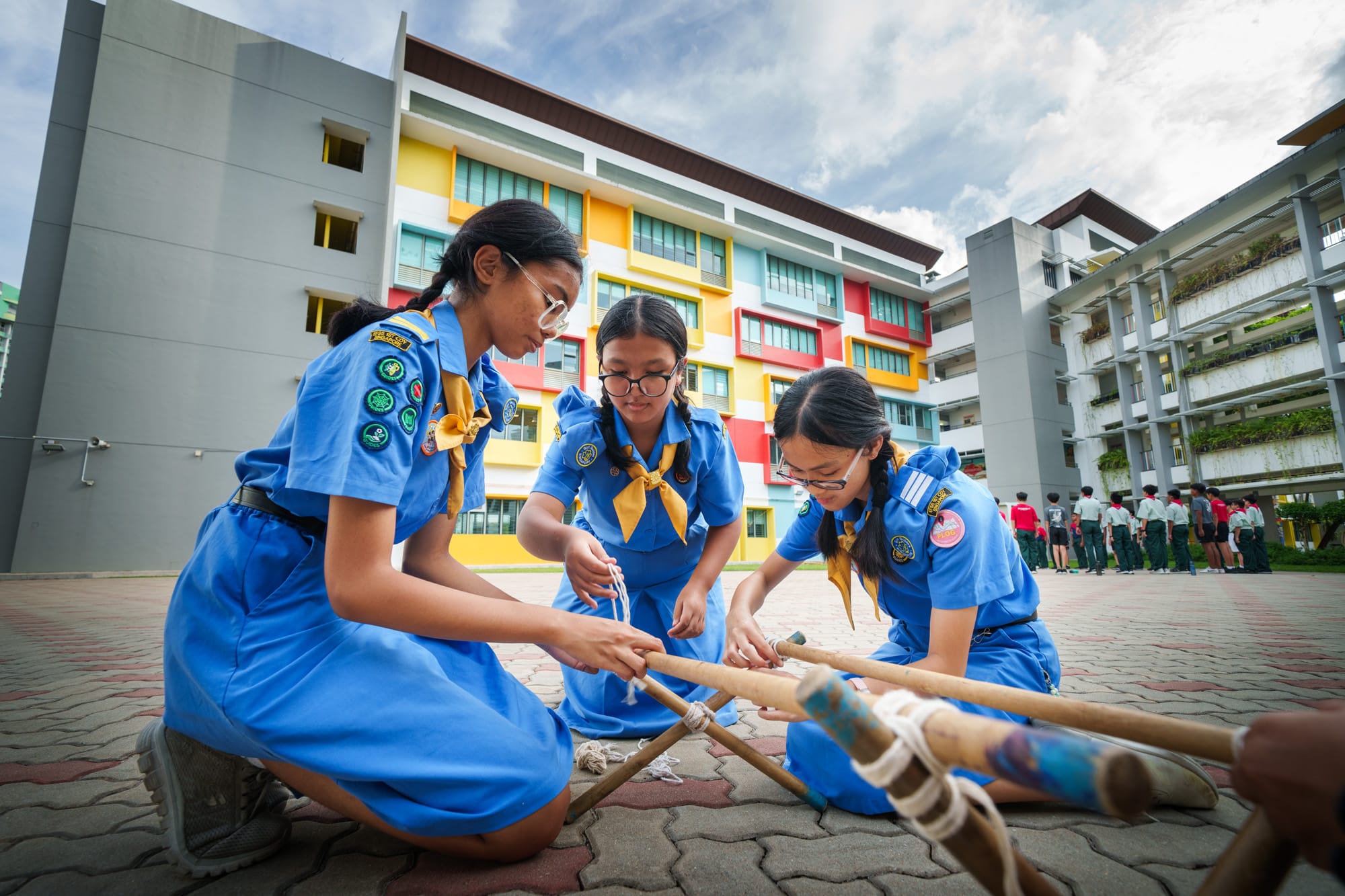 Three girl scouts in blue uniforms kneel outdoors, tying rope around bamboo poles near a colorful school building.