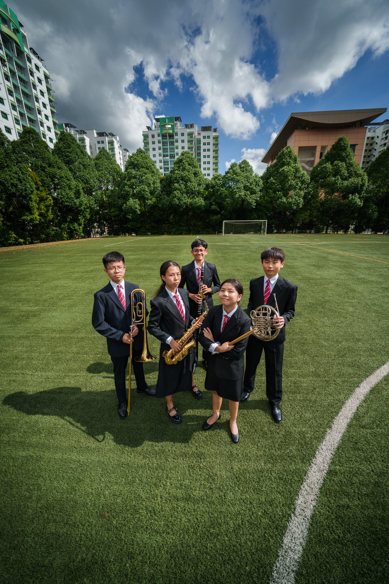 Five students in black uniforms hold brass and woodwind instruments on a green sports field with buildings behind.