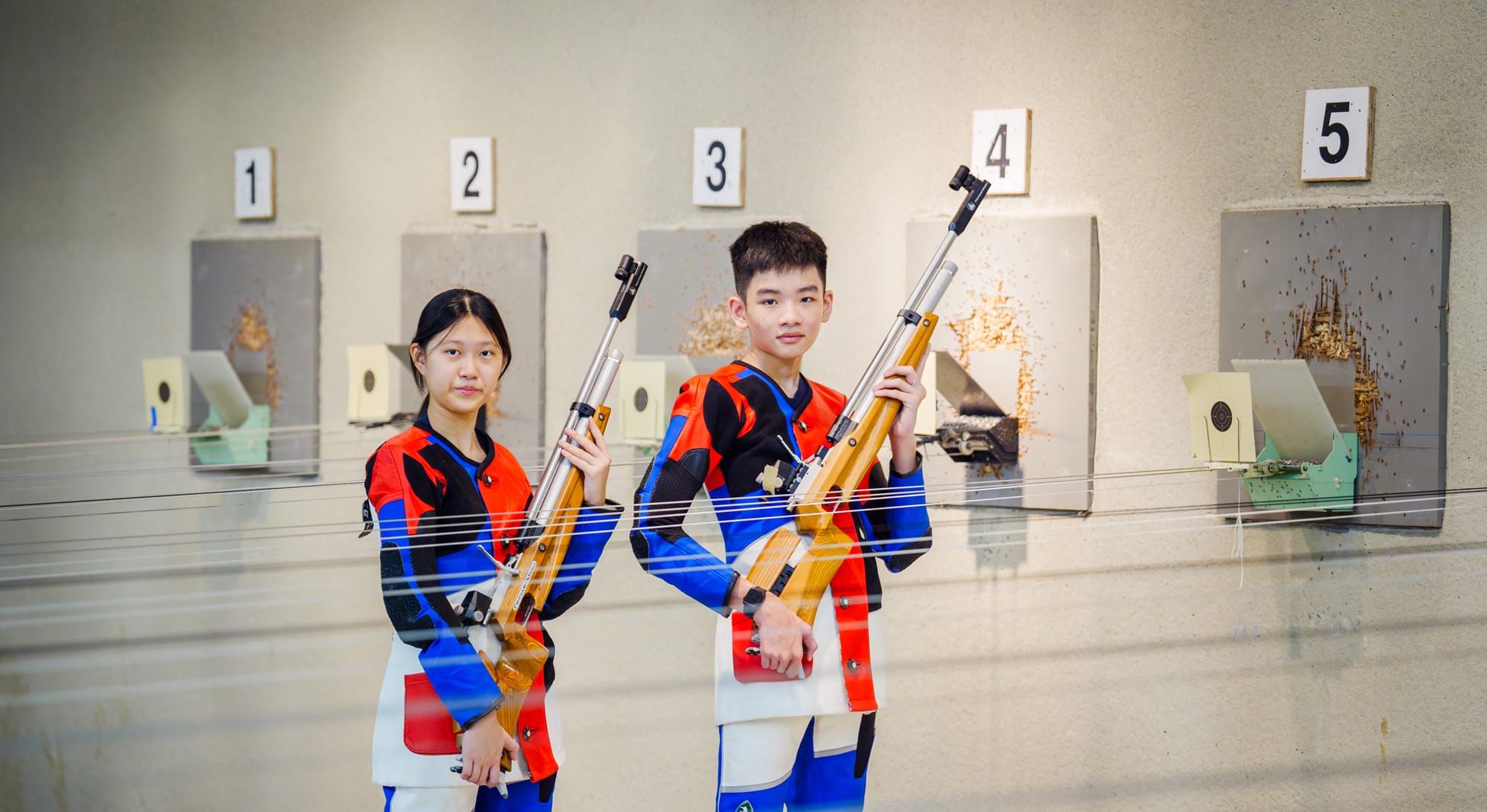Two young shooters in red and blue uniforms hold rifles at an indoor shooting range with numbered target lanes.