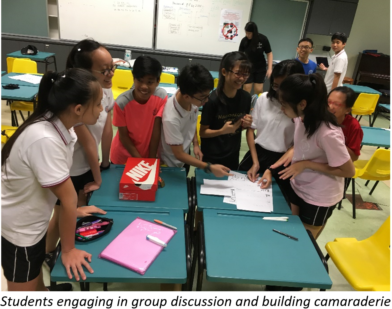 Several students huddle around teal desks with papers and a Nike box in a classroom with a whiteboard.