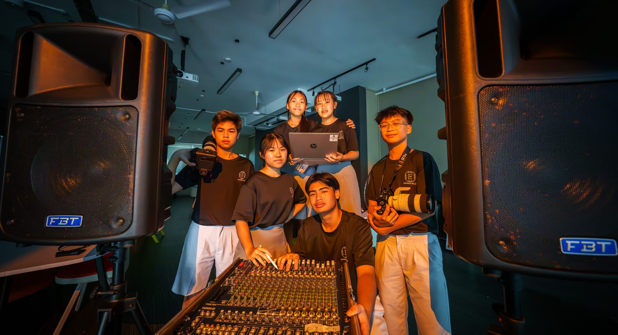Six students pose with audio-visual equipment including FBT speakers, a mixing board, cameras, and a laptop.