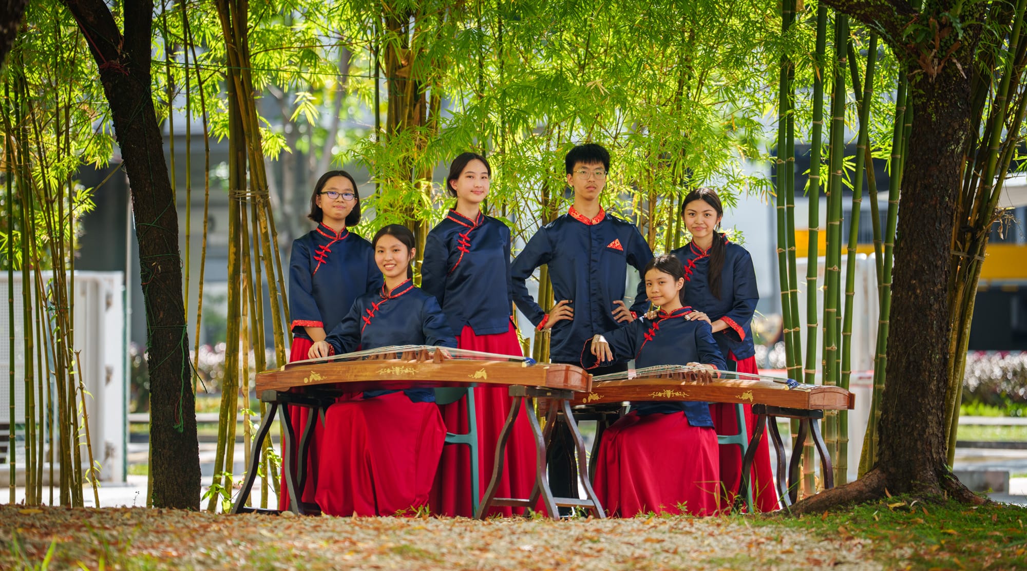 Six young musicians in navy and red traditional Chinese attire pose with guzheng instruments among bamboo trees.