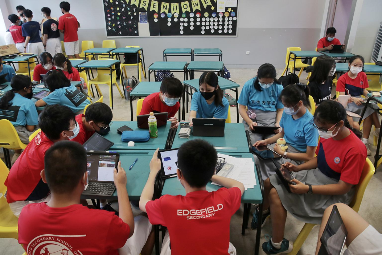 Students in red and blue uniforms wearing masks use tablets at green desks in a classroom.