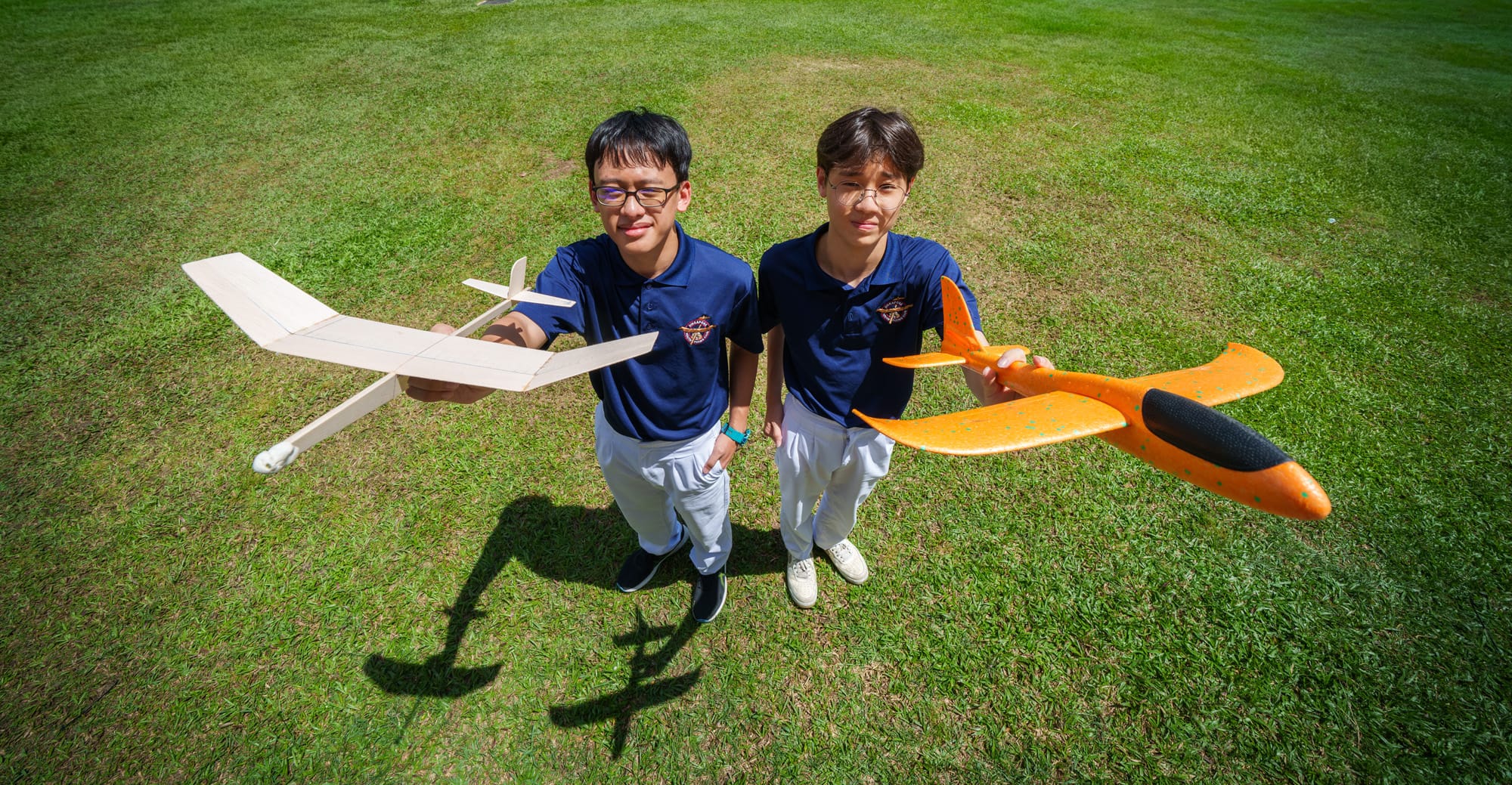 Two boys in navy uniforms stand on grass, one holding a wooden glider and the other an orange foam airplane.