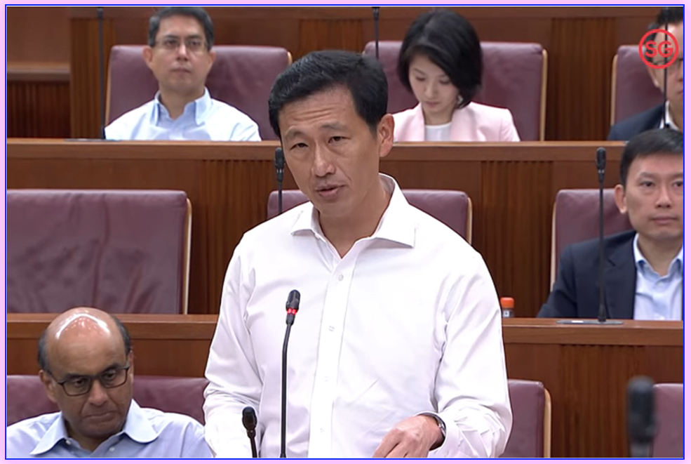 A man in a white shirt speaks at a microphone in a parliament chamber with several seated members behind him.
