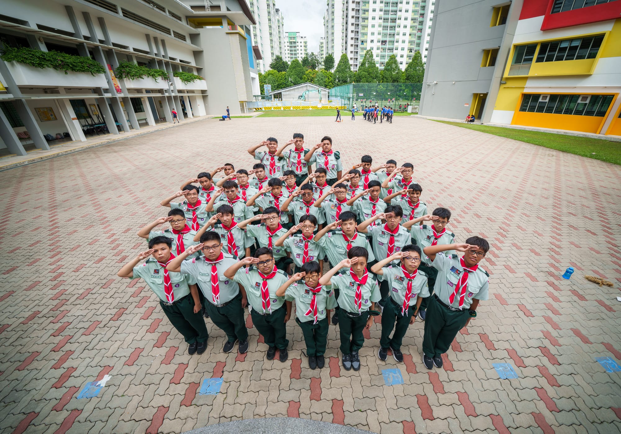 A large group of boys in scout uniforms with red neckerchiefs saluting on a school courtyard.