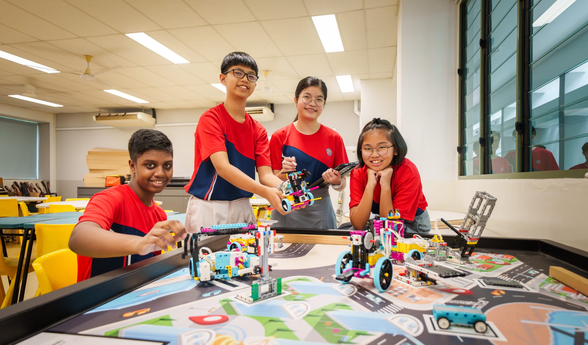 Four students in red uniforms smile around a table with colorful LEGO robots on a map-printed competition mat.