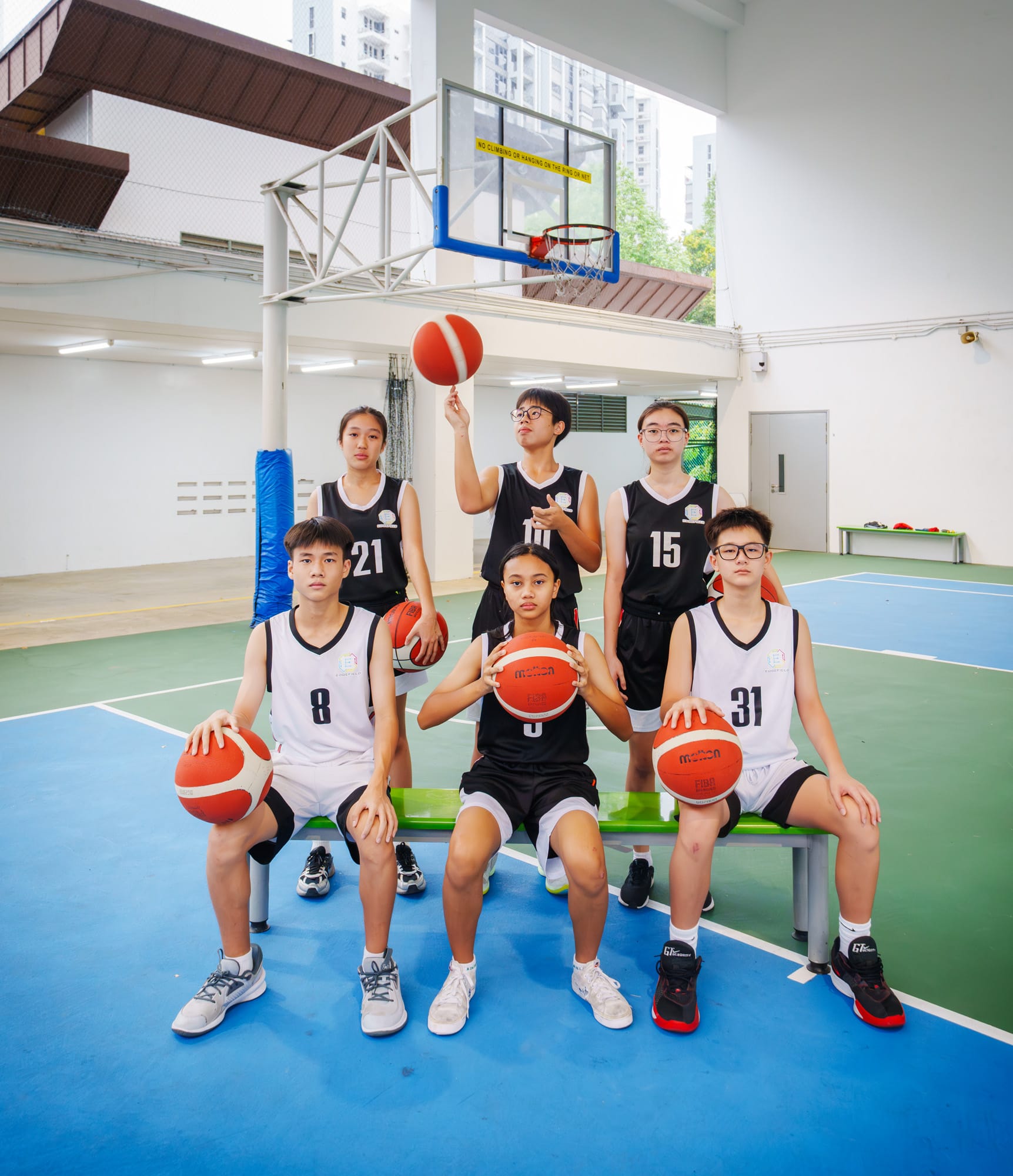 Six young basketball players in black and white jerseys pose on an indoor court, holding balls, with a hoop visible behind them.