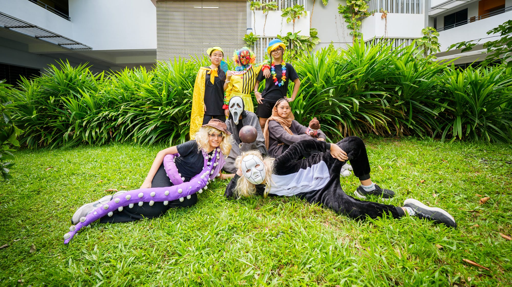Seven people in Halloween costumes including a clown, ghost face mask, and tentacle outfit pose on grass outdoors.