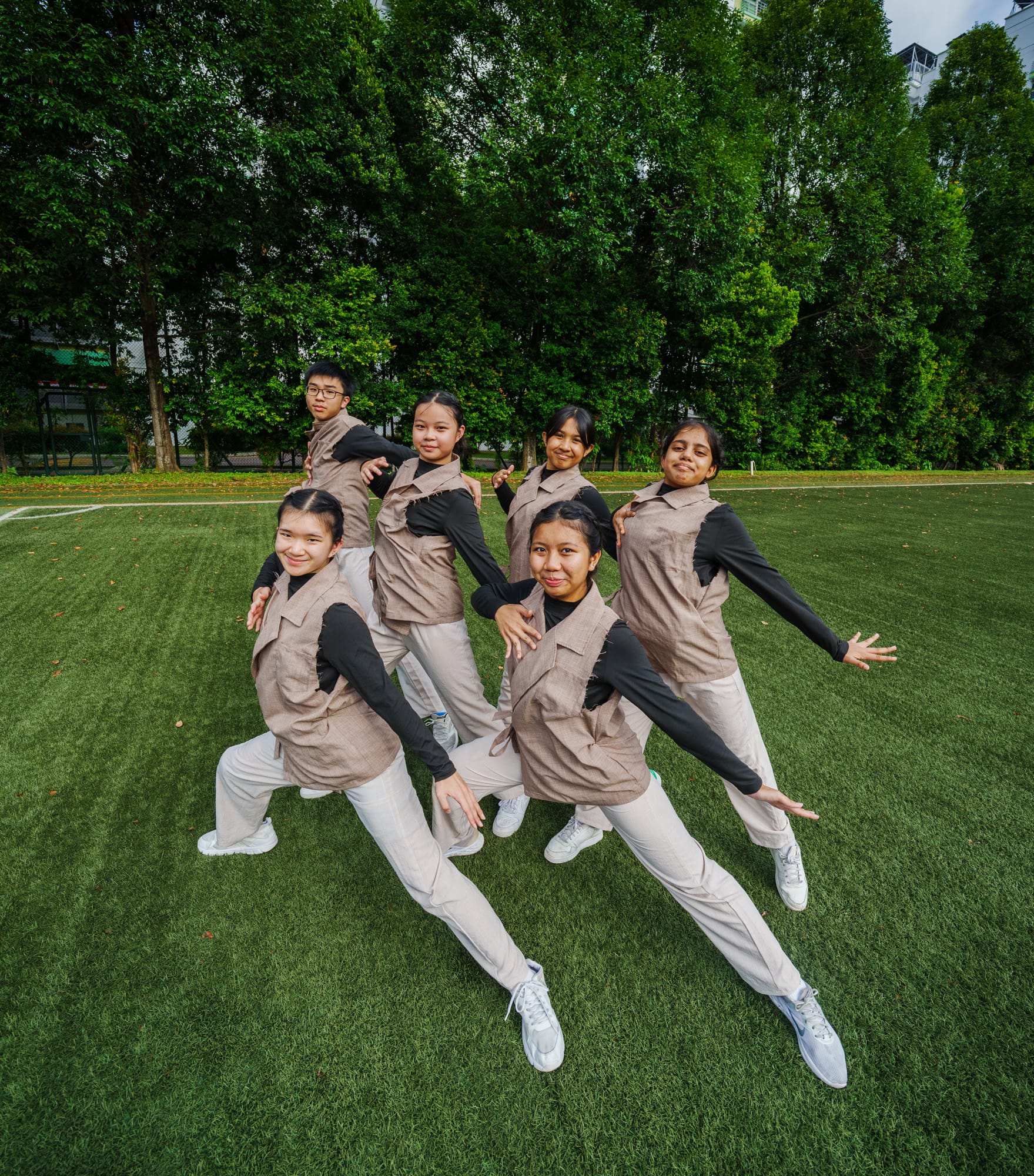 Six youth in matching tan vests and black tops strike dance poses on a green field with trees behind them.