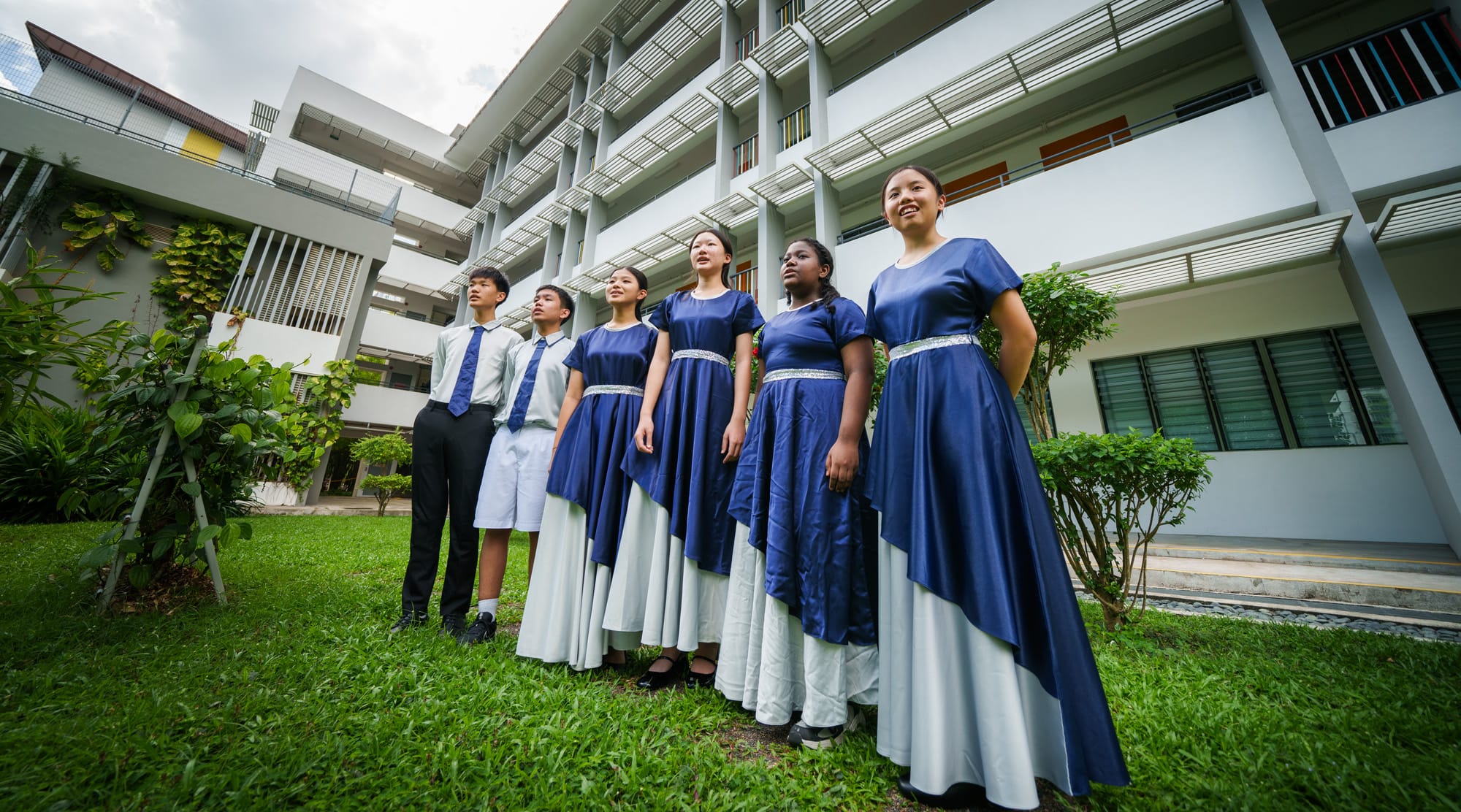 Six students in blue and white choir uniforms stand on grass outside a multi-story school building.