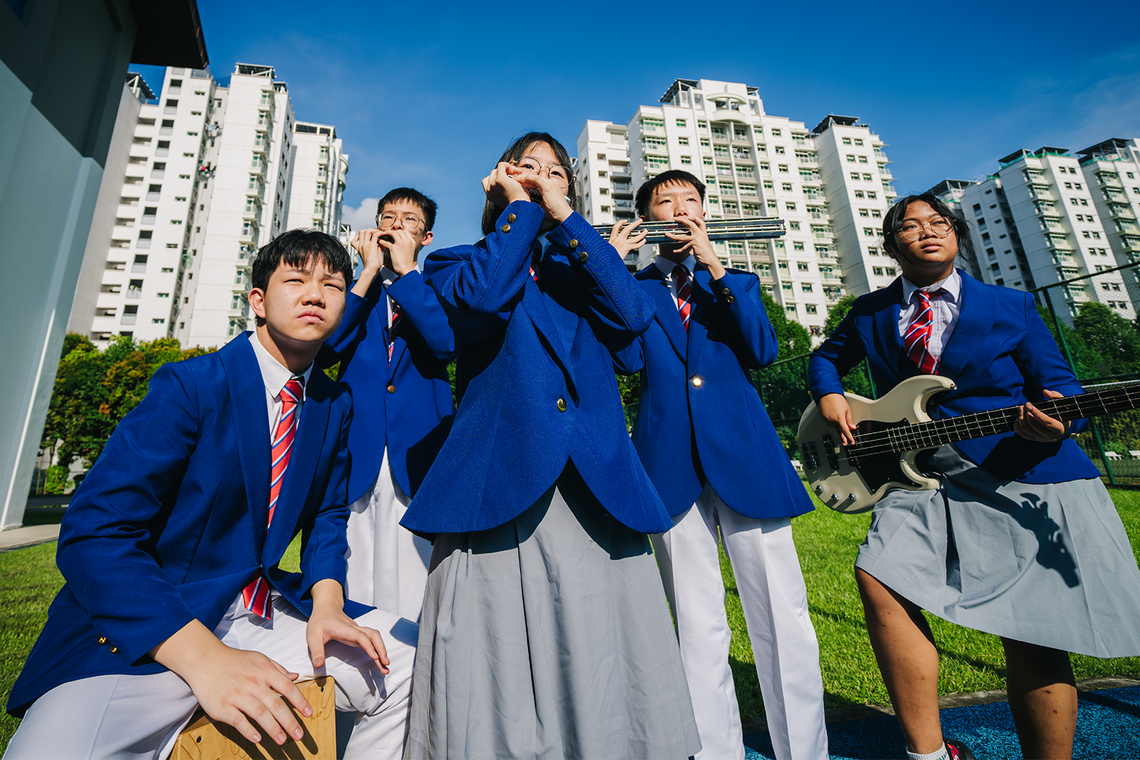 Five students in blue blazers play harmonicas, a guitar, and a cajon outdoors near tall white apartment buildings.