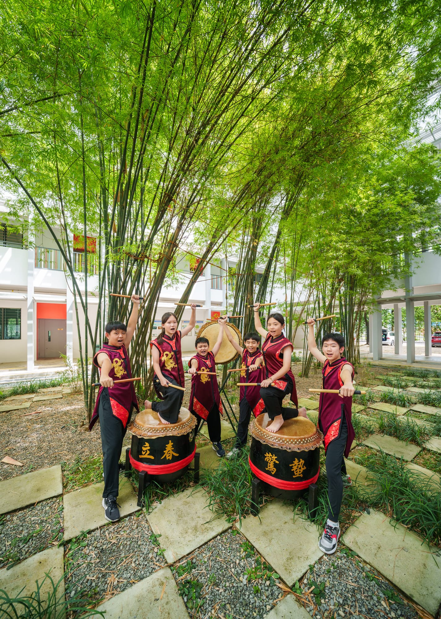 Six children in red and black traditional outfits play large Chinese drums with sticks among bamboo trees outdoors.