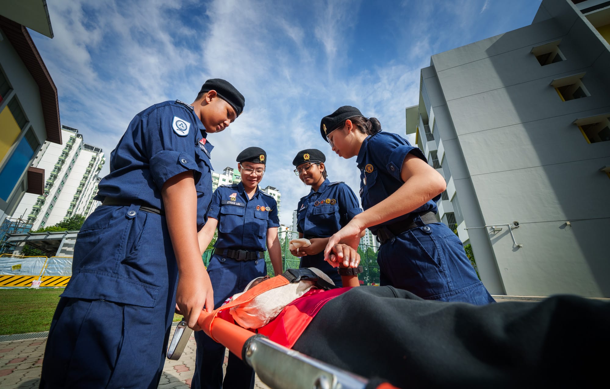 Four young cadets in navy uniforms and black berets attend to a person on a stretcher outdoors near tall buildings.