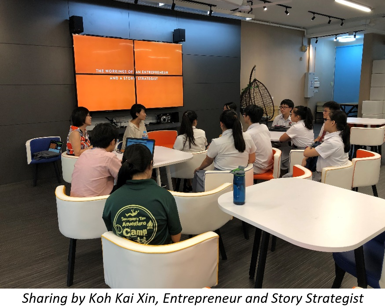 Students sit at tables facing a speaker presenting on a large orange screen about entrepreneurship.