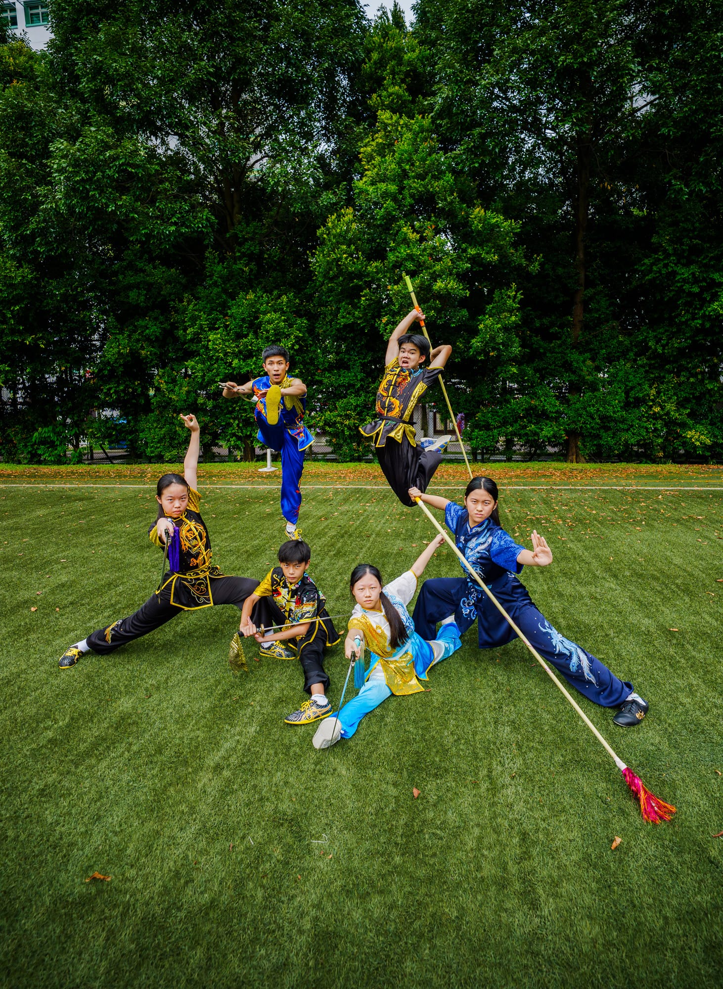 Six young performers in colorful wushu uniforms pose with staffs and swords on a green field with trees behind.