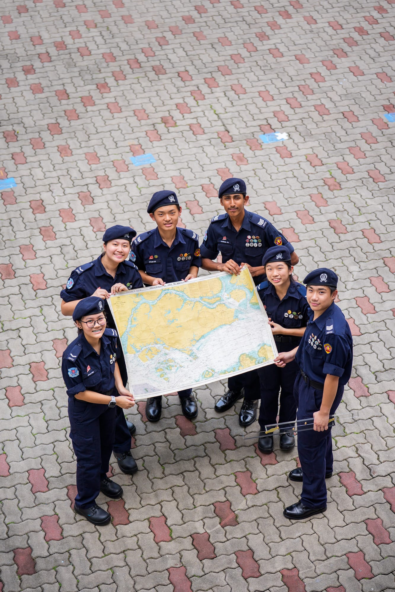 Six young cadets in navy uniforms and berets hold a large nautical map together on a brick courtyard.