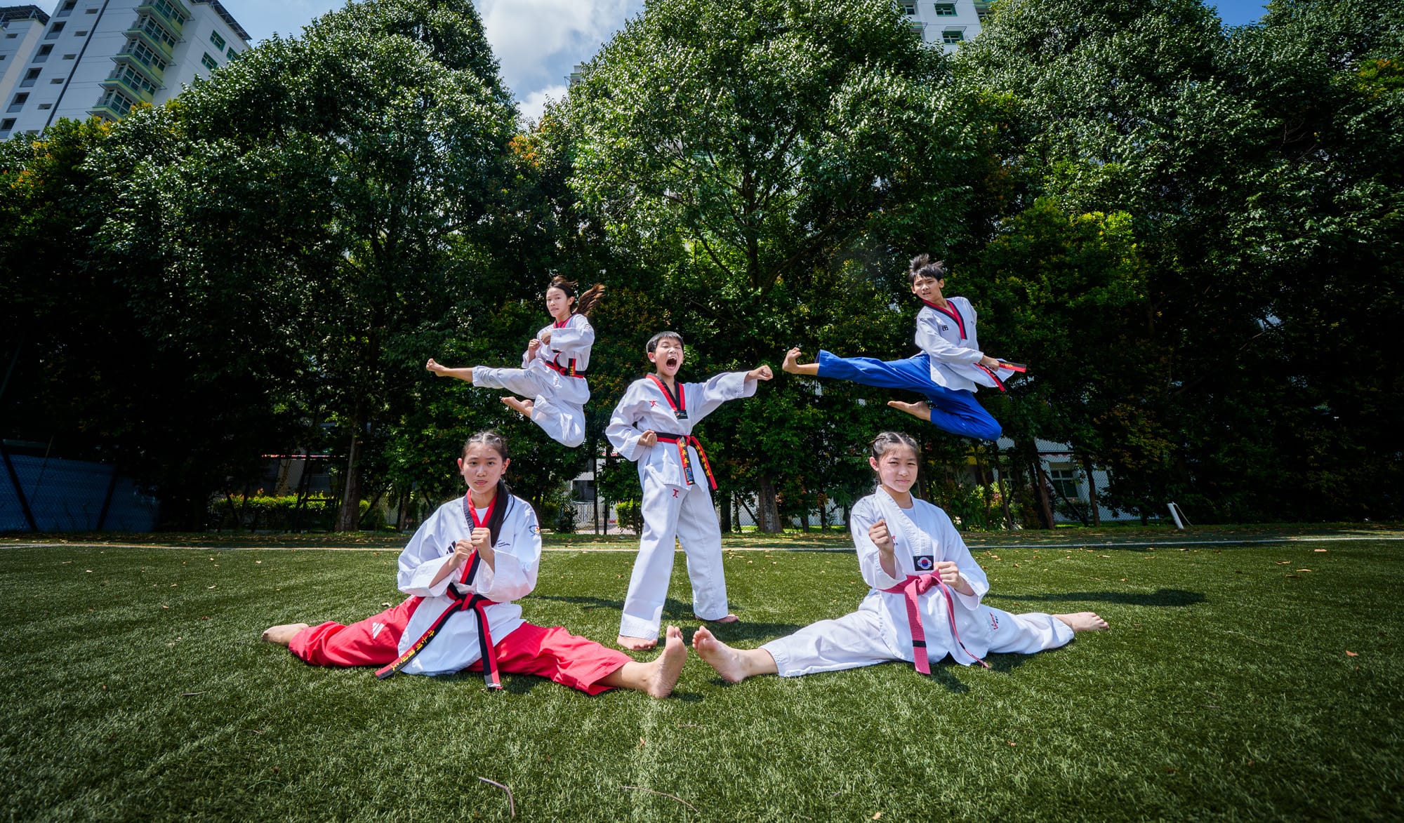Five young taekwondo practitioners in white uniforms pose outdoors, two doing splits and three mid-air kicks on a grass field.